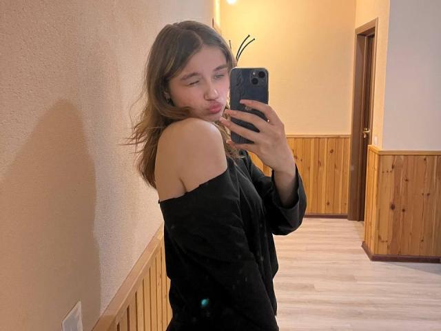 Jenny Osmun, com cabelo castanho claro, vestindo uma blusa preta com um ombro descoberto, tirando uma selfie no espelho em um corredor com painéis de madeira. Ela está fazendo beicinho, com o celular na mão, transmitindo uma aura sexy e confiante..