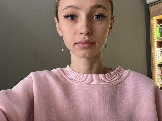 Fotografia ravvicinata di JettaMcClarty, con la pelle chiara, gli occhi azzurri e le lunghe ciglia. Indossa un maglione a coste rosa, piccoli orecchini e ha i capelli castani corti. Illuminazione soffusa, sottile tensione sessuale.