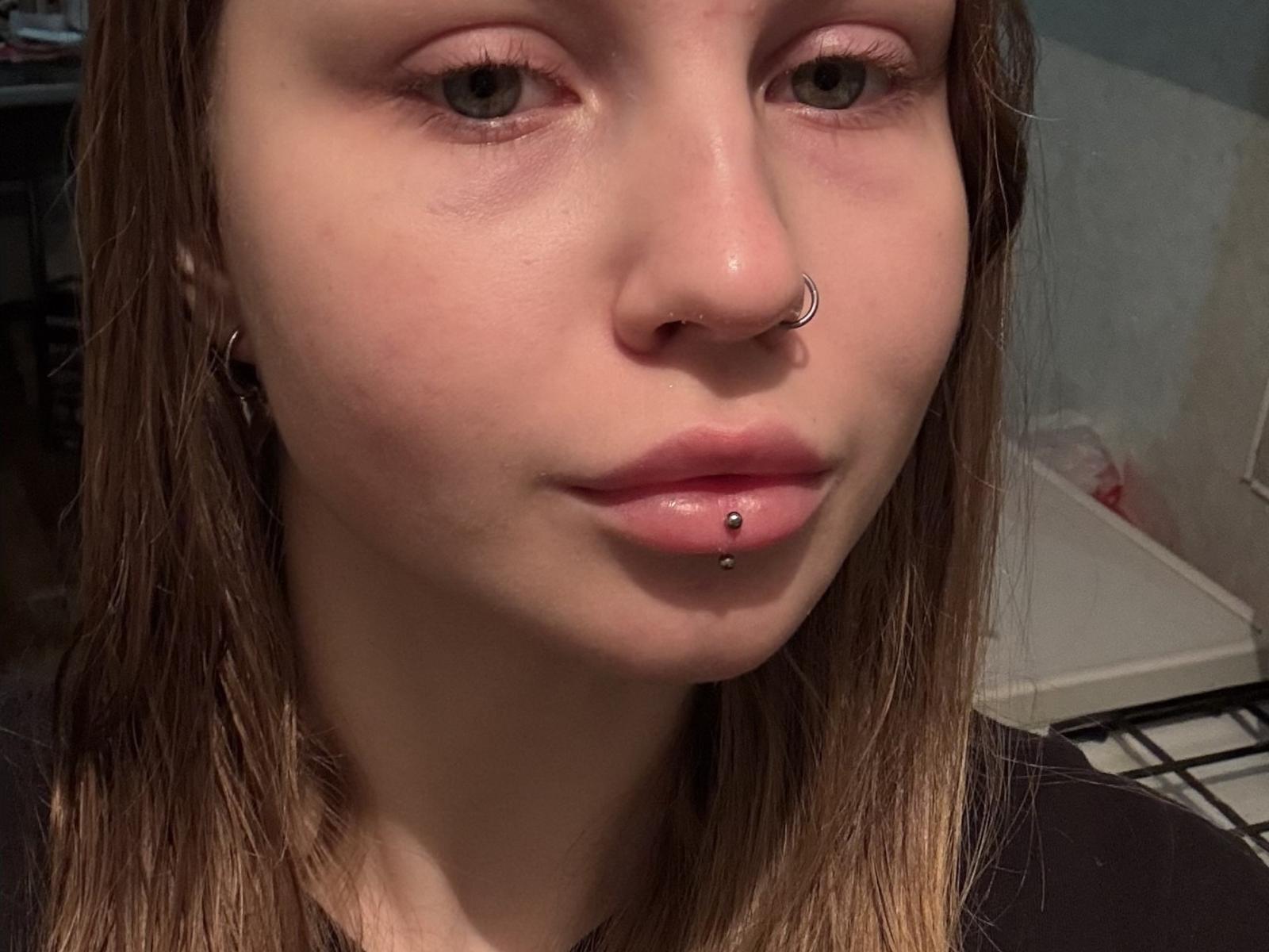 Close-up photo of SimonneRancher, fair-skinned woman with brown hair, nose ring, and lip piercing. Shiny skin, subtle makeup, slight smirk, visible nostrils. Bright light highlights facial features.