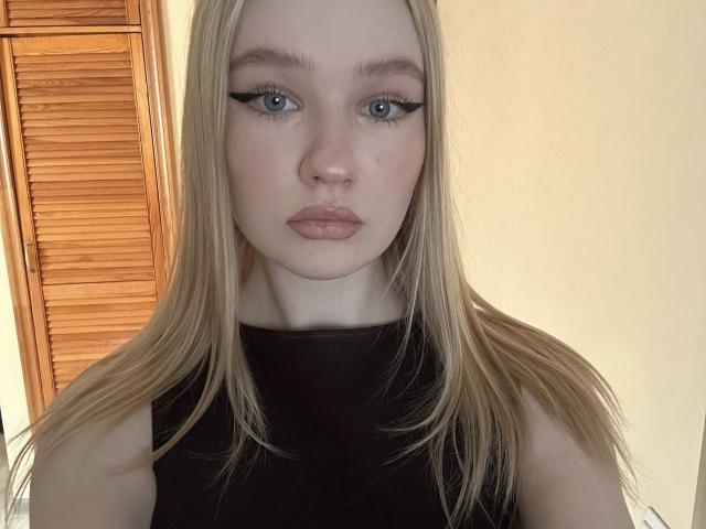 Primo piano di OlympiaCheroki, una donna bionda con occhi azzurri e eyeliner nero, che indossa una canotta nera senza maniche. Ha un'espressione seria, con lunghi capelli e uno sfondo di assi di legno. Molto attraente..