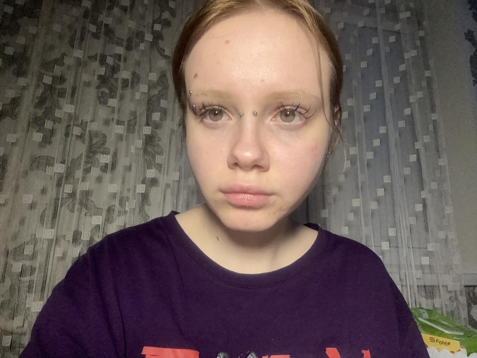 Primer plano de BlytheCooks, de piel clara, con cabello rojo, pecas y labios carnosos, vistiendo una camisa negra. Mirada directa, ligero rubor. El fondo presenta un patrón pixelado en tonos grises. Mirada muy seria..