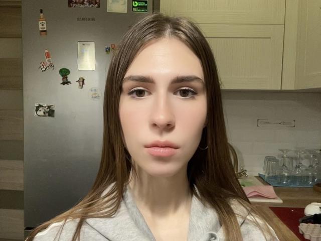 BridgettCatignani with long brown hair, light skin, and brown eyes, wearing a gray shirt, looking directly at the camera with a neutral expression. Her small tits are slightly visible.