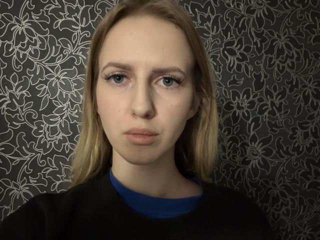 Close-up of LorenaLantey, fair-skinned woman with blue eyes, straight blonde hair, neutral expression, wearing black shirt over blue top. Patterned black background. Focused gaze, slight sexual tension in her look.