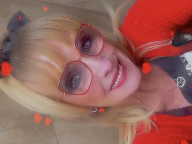 Close-up photo of Jen with blonde hair, red glasses, and a red jacket. She's smiling, showing teeth, with dark purple eyeshadow and red heart effects. Cock is not visible.