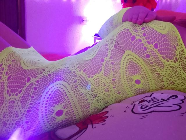 Marriam lying on her side, wearing a yellow fishnet bodysuit that reveals her curvy body and large breasts. Neon purple and white lights glow in the background. Tattoo on her hip.