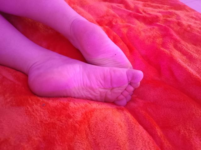 Close-up of Marriam's pale, wrinkled soles and toes, crossed on a vibrant red, textured blanket. Bright pink lighting casts a sensual glow, enhancing the intimate, erotic vibe.
