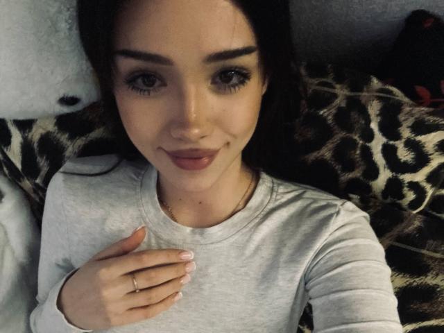 Sherrie Dudycha, sdraiata su una coperta a stampa leopardo, indossa una camicia bianca a maniche lunghe, con una mano sul petto, un leggero sorriso, capelli scuri. occhi, sopracciglia scure, trucco naturale, luce soffusa, selfie casuale, leggera tensione sessuale.