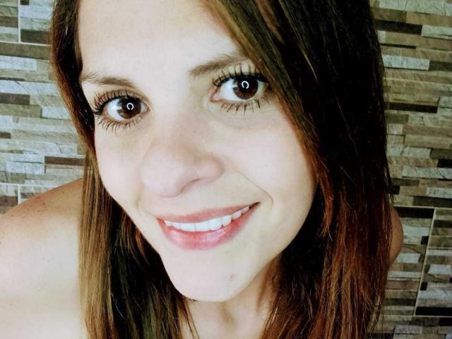 Gros plan d'AbbyySmiith, souriante, avec de grands yeux marron, de longs cheveux bruns et une peau claire. Elle regarde directement l'appareil photo, avec une pointe de tension sexuelle dans son regard. L'arrière-plan présente un motif en carreaux..