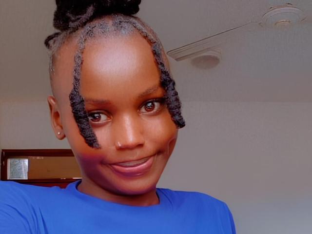 Gros plan de VelvetSavage, avec une peau foncée, des cheveux courts tressés et un sourire confiant. Portant une chemise bleue, souriant directement à la caméra. Visible piercing au nez, éclairage tamisé, pose décontractée..