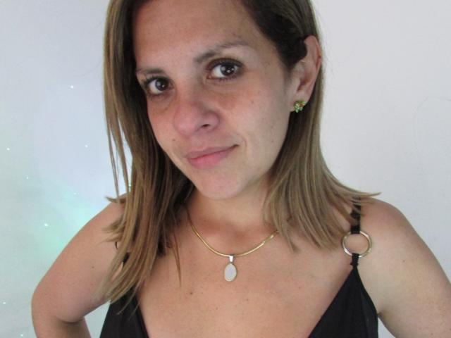 Primo piano di AbbyySmiith con capelli castani di lunghezza media, pelle chiara e occhi marroni. Indossa una maglia nera con un dettaglio dorato a forma di anello, una collana dorata e piccoli orecchini. Un sorriso appena accennato..