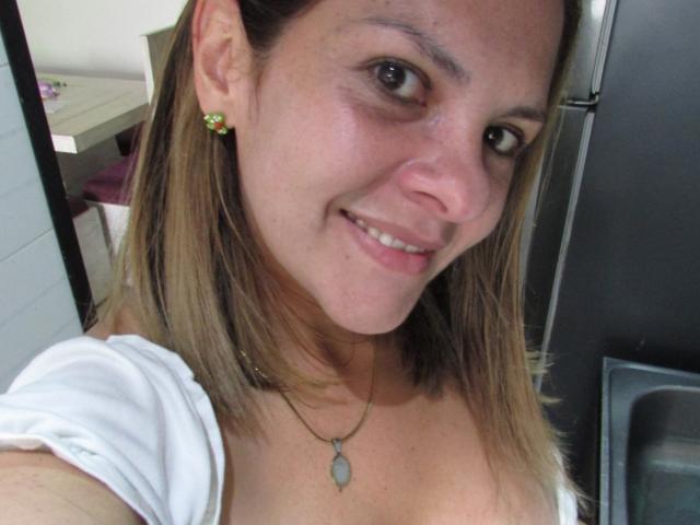 Fotografía de primer plano de AbbyySmiith con cabello castaño claro, sonriendo, vistiendo una blusa blanca, aretes verdes y un colgante. collar. Tiene un aspecto lindo y atractivo, con un escote visible. El fondo incluye un refrigerador..