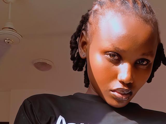 Close-up of VelvetSavage with dark brown skin, braided hair, and intense gaze. She wears a black shirt, leaning forward with sunlight highlighting her face and creating shadows. Her expression is seductive.