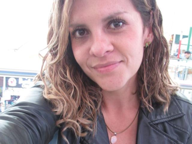 Close-up of AbbyySmiith, a white woman with wavy brown hair, wearing a black leather jacket, smiling slightly. She has brown eyes and a small pink pendant necklace. Background is blurred.