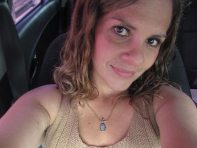 Photo en gros plan d'AbbyySmiith avec des cheveux bruns ondulés, une peau claire et des yeux bleus, portant un débardeur beige et un pendentif. collier, assise dans une voiture, léger sourire, tension sexuelle.