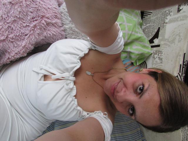 AbbyySmiith, allongée sur le dos, porte une robe blanche moulante avec un décolleté bas, qui expose sa poitrine. Cheveux bruns, yeux bleus, sourire discret. Bras blanc tendu, prenant un autoportrait. Extrêmement sexy..