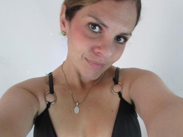 Photo en gros plan d'AbbyySmiith, avec des cheveux bruns courts, une peau claire et de petites boucles d'oreilles. Elle porte un débardeur noir avec des anneaux métalliques sur les bretelles, et elle sourit discrètement. Collier avec pendentif blanc.