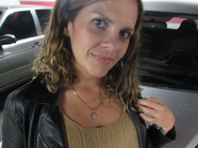 Fotografia di AbbyySmiith con capelli castano corti, che indossa una giacca di pelle nera sopra una maglia beige a maglia. collana con un ciondolo rosa, seduta in macchina, sorride in modo seducente..