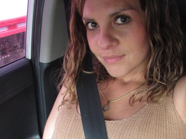 Nahaufnahme von AbbyySmiith mit lockigem, braunem Haar, die ein beiges Strickoberteil und einen schwarzen Sicherheitsgurt trägt und in einem Auto sitzt. Ihr Ausdruck ist leicht verführerisch, mit einem kaum merklichen Lächeln. Rotes Autoscheinwerfer draußen..