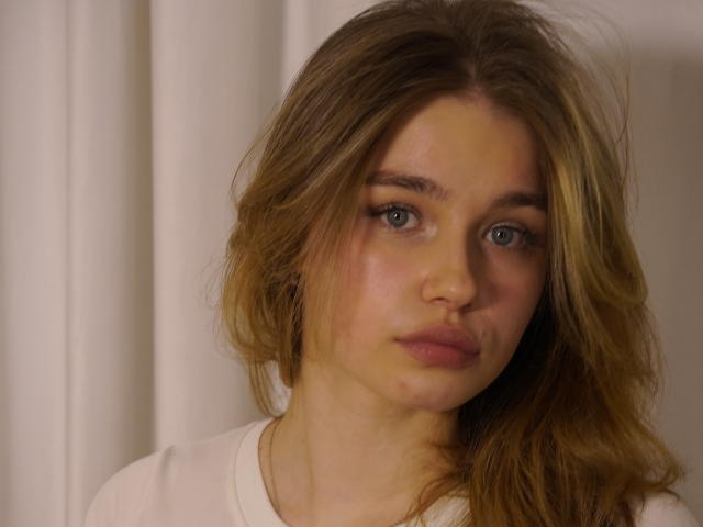 Fotografía de primer plano de JennyOsmun, una joven con ojos azules, cabello ondulado de color castaño claro y mejillas rosadas. Ella lleva una blusa blanca y mira directamente a la cámara con una expresión ligeramente seria..