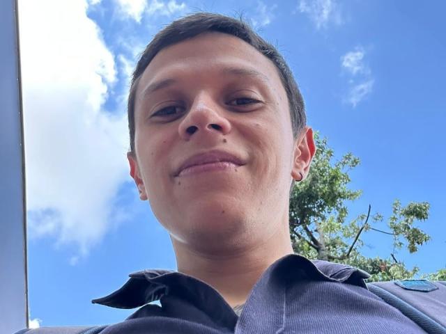 Close-up foto van MasterDaveExtreme, die naar beneden kijkt naar de camera met een lichte grijns. Blauwe lucht en witte wolken op de achtergrond, een groene boom aan de rechterkant. Het dragen van een donker shirt..