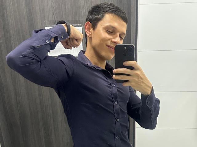 MasterDaveExtreme, a handsome guy with short black hair, wearing a tight navy blue shirt, takes a selfie with a confident smirk. He raises his arm, flexing his bicep, showing off his fit physique.