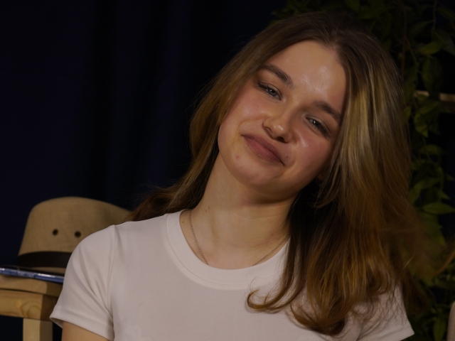 Fotografie von Jenny Osmun mit schulterlangem, braunem Haar, die leicht lächelt und ein weißes T-Shirt trägt, stehend vor einem dunkler Hintergrund mit einem Hut auf einem Tisch auf der linken Seite. Sie strahlt eine subtile sexuelle Spannung aus..