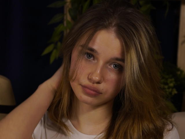 Nahaufnahme eines Fotos von JennyOsmun, einer jungen Frau mit heller Haut, blauen Augen und braunen Haaren, die direkt in die Kamera schaut. eine Kamera, auf der man ihr eine leichte Röte auf den Wangen und eine Hand in ihrem Haar sieht. Die sexuelle Spannung ist spürbar..