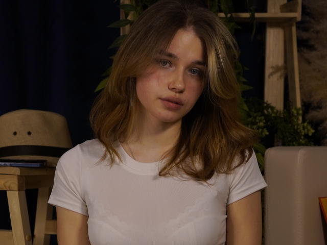 Photographie de JennyOsmun, avec des cheveux bruns de longueur moyenne, portant un t-shirt blanc, assise dans une pièce peu éclairée. Elle a un visage légèrement rougi, un regard doux et une tension sexuelle visible..