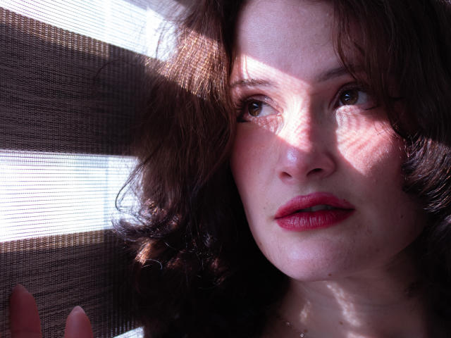 Close-up de DanaStone, uma mulher atraente com cabelo castanho encaracolado, lábios vermelhos e pele radiante. A luz forte cria sombras intensas em seu rosto. Seu olhar é direto, com um ar sedutor..