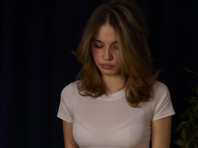 Jenny Osmun, met bruin haar, draagt een strak wit T-shirt dat haar kleine borsten accentueert. Ze kijkt naar beneden met een verleidelijke uitdrukking, donkere achtergrond, subtiele schaduwen, zachte belichting..