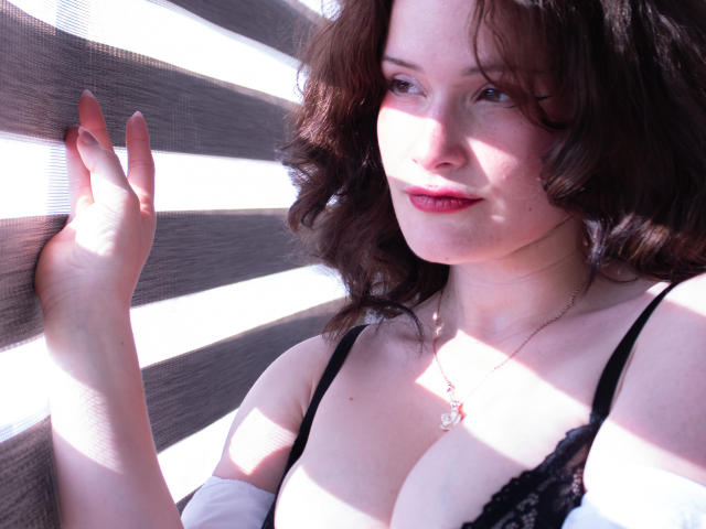 Close-up photo of DanaStone with shoulder-length brown hair, wearing a black lace bra, illuminated by bright white light beams. DanaStone's hand rests against the wall, eyes slightly closed, smiling.