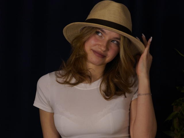 Jenny Osmun lleva un sombrero de paja color beige con una banda negra, sonriendo sutilmente y tocándose el sombrero con la mano derecha. Ella lleva una blusa blanca, ligeramente transparente, sobre un fondo oscuro..