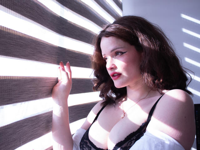 DanaStone with dark brown hair, wearing a black lace bra, illuminated by bright white lights. Her hand touches the wall, casting shadows on her face. Bright light, dark shadows, intense gaze.