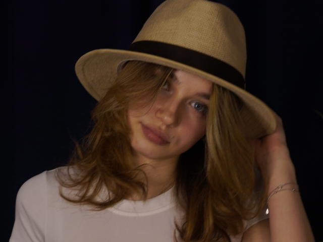JennyOsmun, met bruin haar, draagt een beige fedora met een zwarte band, een wit T-shirt, op een donkere achtergrond, lichtjes gekanteld. Hoofd, hand die hoed aanraakt, blauwe ogen, subtiele glimlach, armbandje om de rechterpols. Heel schattig.
