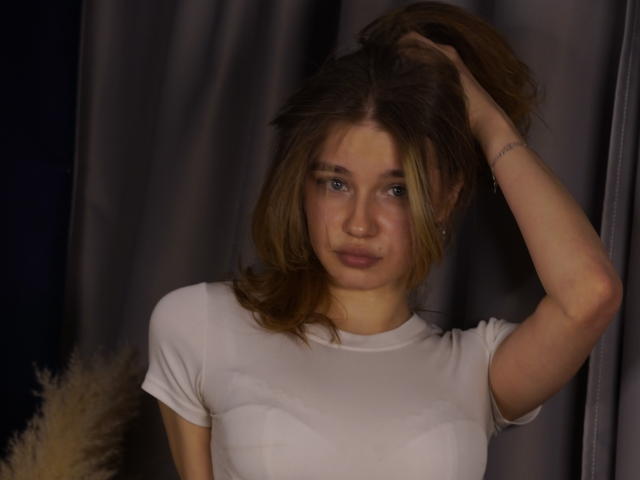 Jenny Osmun, con i capelli castano disordinati, una mano legata in una coda di cavallo, indossa una maglietta bianca aderente, ha gli occhi azzurri, un leggero sorriso e un incarnato chiaro. illuminazione, sfondo con tenda grigia, ombre morbide, casual, sexy, espressione leggermente infastidita..