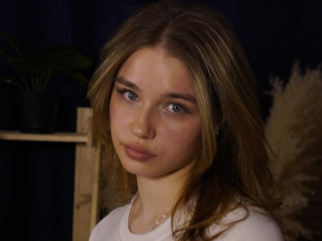 Photographie en gros plan de JennyOsmun, une jeune femme à la peau claire, aux yeux bleus et aux cheveux bruns, portant une chemise blanche et regardant droit devant elle. Directement face à la caméra, avec une expression subtile et séduisante. Fond noir.