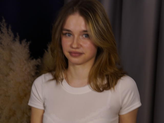 Jenny Osmun hat hellbraunes, schulterlanges Haar, blaue Augen und ein leichtes Lächeln. Sie trägt ein enges, weißes T-Shirt, das ihre kleinen Brüste betont. Der Hintergrund ist dunkel und hat eine weiche, flaumige Textur..