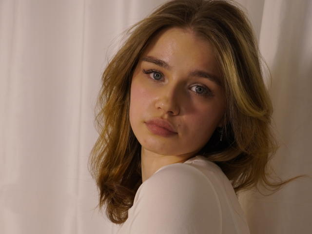 Fotografia em close-up de JennyOsmun, uma jovem com cabelos castanhos ondulados, olhos azuis e um rosto levemente avermelhado. Ela veste uma blusa branca, olhando diretamente para a câmera com uma expressão sutil e sedutora..