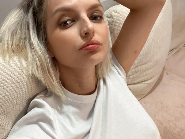 Close-up photo of DouceAmelia, blonde hair, light skin, wearing a white t-shirt, reclining on a beige couch, arm raised behind head, intense gaze, slight smirk, soft natural light, relaxed pose.