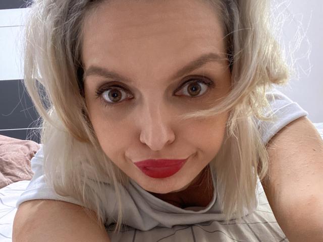 Close-up foto van DouceAmelia, blond, blauwe ogen, rode lippenstift, voorovergebogen, witte mouwloze top, intense blik. licht rommelig haar, natuurlijk licht, witte achtergrond, lichte schaduw, suggestieve uitdrukking.