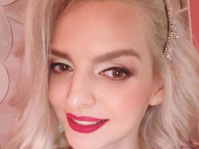 Primo piano di DouceAmelia con capelli biondo platino, che indossa rossetto rosso e un copricapo scintillante. Sorride in modo seducente, con un trucco per gli occhi molto marcato e occhi marrone scuro. Le sue labbra sono leggermente socchiuse.