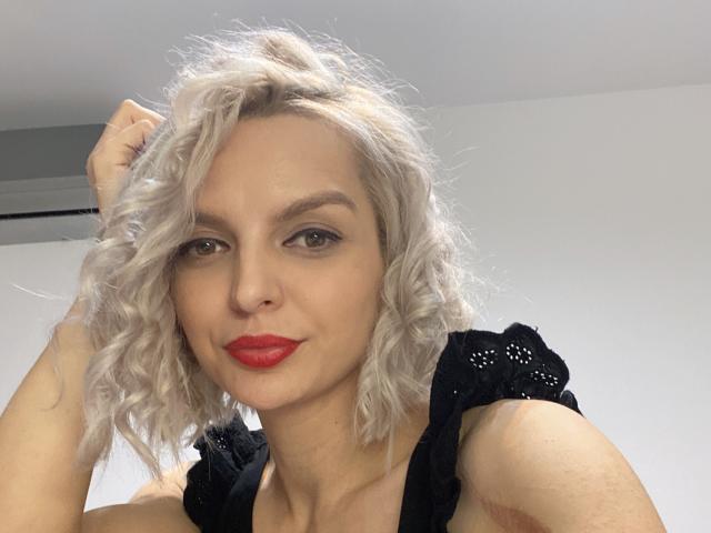 Gros plan de DouceAmelia, une femme blonde aux cheveux bouclés, au rouge à lèvres rouge et portant un haut noir, avec une main dans les cheveux, peau claire, et un regard séduisant. expression, éclairage doux, fond blanc, légère ombre sur le visage.