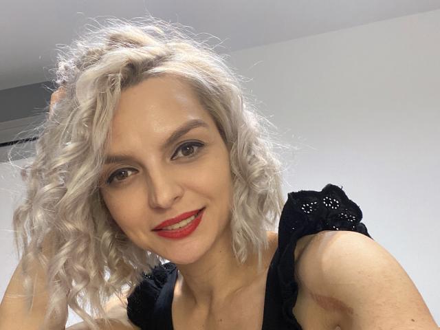 Autoportrait en gros plan de DouceAmelia, une femme blonde aux cheveux bouclés, au rouge à lèvres rouge et portant un haut noir, regardant directement l'objectif. appareil photo, avec un léger sourire, dégageant une tension sexuelle. L'éclairage vif met en valeur ses traits..