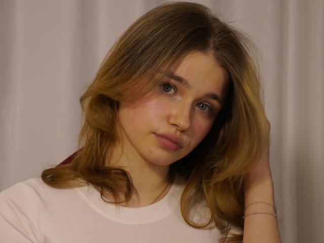 Fotografía de JennyOsmun con cabello castaño hasta los hombros, vistiendo una camisa blanca, mirando directamente a la cámara con cabeza ligeramente inclinada y una expresión neutra. Fondo de color gris claro. Iluminación suave..