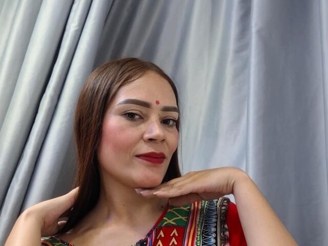Photographie de ShantalaKrisha, une femme aux cheveux bruns, avec un point rouge sur le front, du rouge à lèvres rouge et une robe traditionnelle colorée. Elle pose sa main sur sa joue, avec des rideaux gris en arrière-plan. Extrêmement sexy..