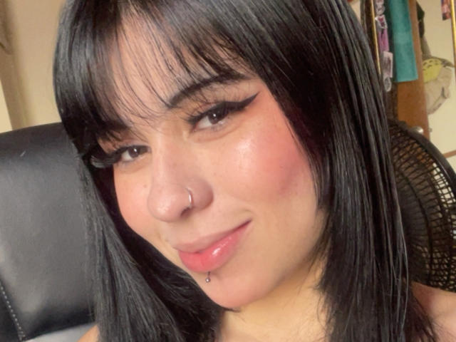 Close-up foto van GaiaJoness, een jonge vrouw met recht zwart haar, een pony en een neusring. Ze heeft een lichtroze huid, donkere eyeliner, een lip-piercing en een subtiele glimlach. Achtergrond wazig.