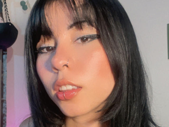 Close-up of GaiaJoness with long black hair, heavy black eyeliner, and glossy red lips. GaiaJoness has a slight tongue piercing visible. Fucking sexy look, dark eyes, light skin.