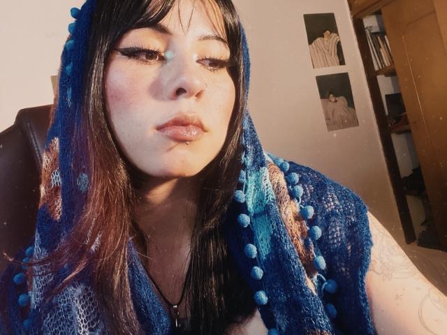 Close-up de GaiaJoness, uma jovem com cabelos longos e pretos, usando um capuz azul com contas, olhando diretamente para a câmera. Com um visual de lábios levemente entreabertos e com brilho. A luz do sol realça seu rosto..