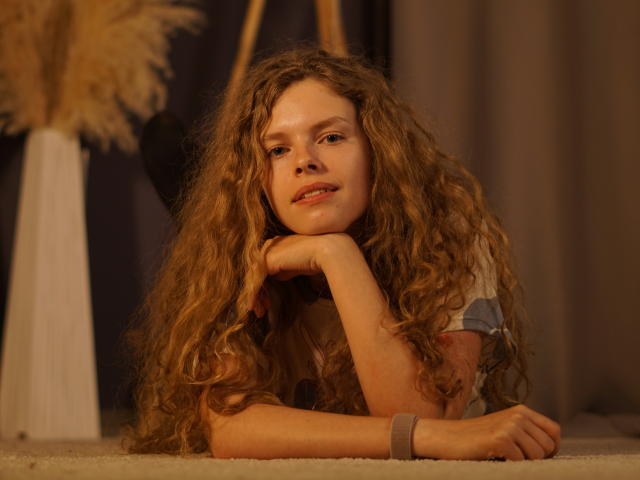 TamekiaIafrate lies on carpeted floor, head resting on hand, long curly brown hair cascading over shoulders. Soft, warm lighting, relaxed expression, slightly parted lips, bracelet on wrist. Dimly lit background.