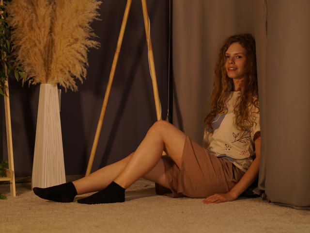 Tamekia Iafrate zit tegen een grijze gordijn aan, met gespreide benen, en draagt een beige rok, zwarte sokken en een witte top. Haar lange, krullende haar en stralende gezicht creëren een sexy, ontspannen sfeer. Vaas met pampa gras op de achtergrond.